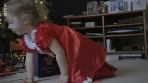 Une jeune enfant portant une robe rouge de Noël joue au sol devant un sac cadeau portant l'inscription Buckingham Palace, dans un intérieur décoré pour les fêtes de fin d'année.