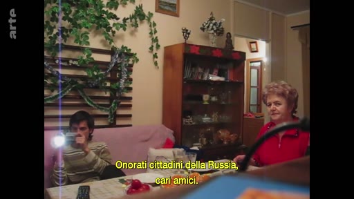 Une femme et un jeune homme filmant avec une caméra lors d'une célébration du réveillon du Nouvel An à Marioupol. Le sous-titre en italien indique 'Onorati cittadini della Russia, cari amici'.