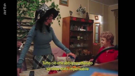 Une jeune femme portant un chapeau festif et une femme âgée en pull rouge sont assises à une table dans un intérieur domestique à Marioupol, lors d'une célébration du Nouvel An. Une inscription en italien apparaît à l'écran, soulignant le caractère inattendu de la situation.