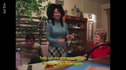 Une famille ukrainienne réunie autour d'une table pour célébrer le Nouvel An à Marioupol, avec des sous-titres en italien affichés à l'écran. La scène montre une atmosphère domestique intime dans un intérieur privé.