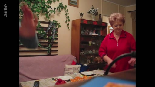 Une femme d'âge mûr, vêtue d'un haut rouge, s'affaire à préparer une table dans une salle à manger décorée de plantes vertes, dans un contexte de vie quotidienne.
