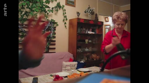 Une femme d'âge mûr en haut rouge prépare à manger dans une cuisine, avec une main floue au premier plan. Cette scène illustre la vie quotidienne dans un intérieur privé.