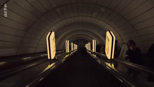 Vue en perspective d'un long escalator dans une station de métro souterraine, utilisée comme abri par des civils. L'éclairage tamisé souligne l'architecture voûtée du tunnel.
