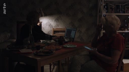 Une famille ukrainienne est réunie autour d'une table pour un repas en soirée dans un intérieur domestique sombre. Une personne utilise un ordinateur portable tandis qu'une autre consulte un téléphone mobile, illustrant le quotidien en Ukraine.