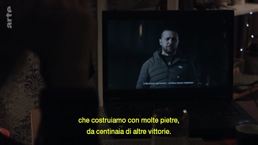 Le président ukrainien Volodymyr Zelensky apparaît sur l'écran d'un ordinateur portable dans un intérieur domestique, accompagné de sous-titres en italien. Cette image illustre la diffusion de messages officiels du chef d'État ukrainien pendant le conflit en Ukraine.