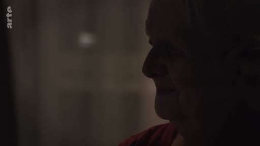 Gros plan sur le visage d'une femme âgée dans une pièce sombre, illustrant le quotidien d'un foyer ukrainien en soirée.