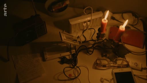 Une vue en plongée d'une table encombrée d'objets du quotidien, incluant des bougies allumées, un téléphone portable, des lunettes et une radio, évoquant une situation de coupure d'électricité ou de précarité.