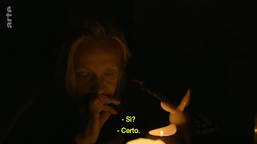 Une femme âgée, filmée en gros plan dans une ambiance sombre et intimiste, est éclairée par une lampe décorative en forme de lapin. Elle semble engagée dans une conversation, comme l'indiquent les sous-titres à l'écran.