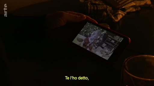 Une personne tient un smartphone dans une pièce sombre, visionnant une vidéo montrant une scène de vie quotidienne ou d'archive. L'image est accompagnée d'un sous-titre en italien.