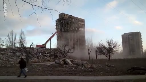 Une pelleteuse démolit un immeuble d'habitation gravement endommagé à Marioupol, en Ukraine, dans le cadre des opérations de nettoyage après les combats. La scène montre les vestiges de la ville et les travaux de démolition en cours.