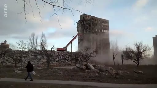 Une pelleteuse démolit un immeuble résidentiel gravement endommagé à Marioupol, en Ukraine, dans un contexte de guerre. Un piéton marche au premier plan devant les décombres et la poussière soulevée par les travaux de démolition.