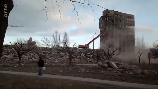Vue d'ensemble d'une opération de démolition d'un immeuble résidentiel gravement endommagé à Marioupol, en Ukraine. Une pelleteuse est visible en train de détruire la structure tandis qu'un piéton marche à proximité dans un paysage urbain dévasté.
