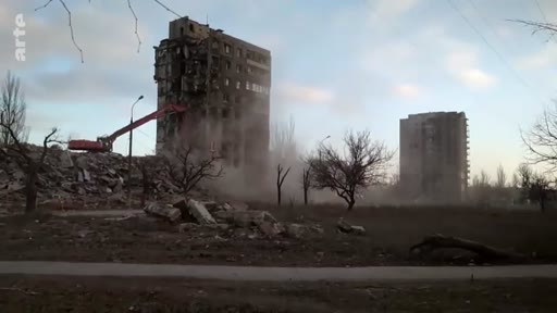 Une pelleteuse à bras long procède à la démolition d'un immeuble d'habitation gravement endommagé à Marioupol, en Ukraine. Cette scène illustre les opérations de nettoyage et de reconstruction urbaine dans une zone marquée par les combats.
