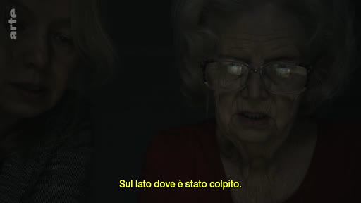 Une femme âgée portant des lunettes est filmée en gros plan lors d'un entretien, avec une autre personne partiellement visible à ses côtés. Le sous-titre indique 'Sul lato dove è stato colpito'.