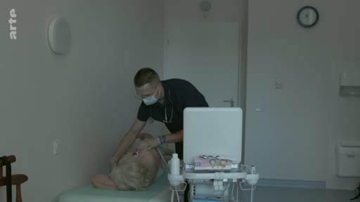 Un médecin portant un masque chirurgical effectue un examen médical sur un patient âgé allongé sur une table dans un cabinet médical. La scène illustre une consultation médicale de routine ou un suivi de santé.