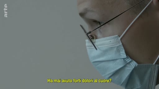 Un médecin portant un masque chirurgical et des lunettes interroge un patient lors d'une consultation médicale. Le sous-titre à l'écran demande : 'Ha mai avuto forti dolori al cuore?'