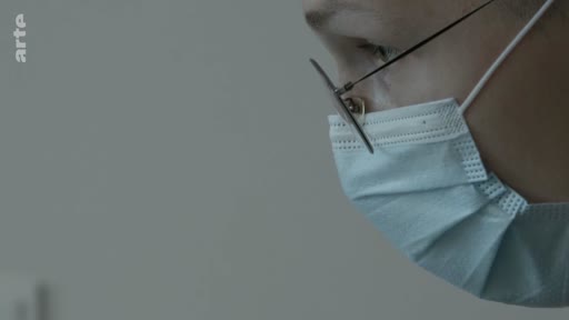 Gros plan sur le visage d'un médecin portant un masque chirurgical et des lunettes, dans un contexte de soins médicaux ou de pandémie.