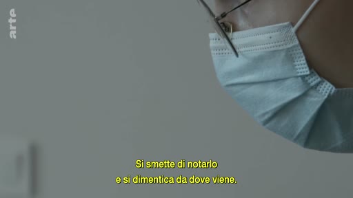 Gros plan sur le visage d'une personne portant un masque chirurgical et des lunettes, accompagnée d'un sous-titre en italien sur la perception et l'oubli.