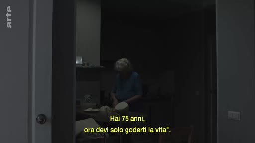 Une femme âgée de 75 ans est filmée dans sa cuisine à Londres, illustrant son quotidien. Le sous-titre italien souligne l'importance de profiter de la vie à cet âge.