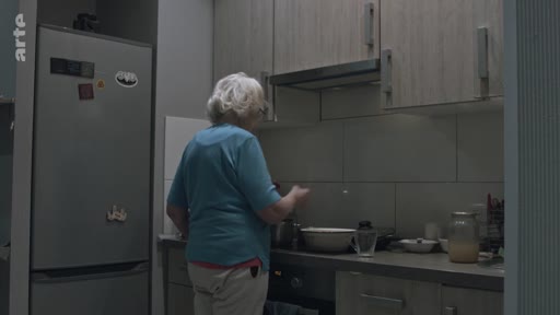Une femme âgée aux cheveux blancs est vue de dos en train de cuisiner dans une cuisine domestique, illustrant le quotidien d'une personne âgée dans le cadre d'un reportage sur les difficultés familiales à Londres.