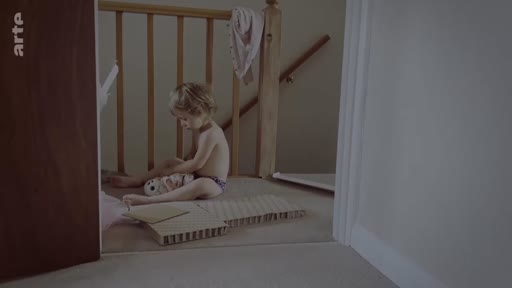 Un jeune enfant, en couche, est assis sur la moquette d'un palier près d'une rampe d'escalier en bois, jouant avec des objets dans une maison à Londres.