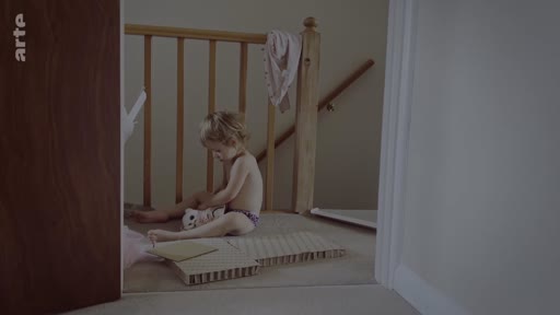 Un jeune enfant en couche-culotte joue avec un petit jouet sur le sol d'un palier près d'une rampe d'escalier dans une maison à Londres. Cette scène illustre le quotidien et l'intimité familiale dans le cadre d'un reportage sur les difficultés rencontrées par une famille.