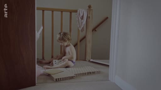 Un jeune enfant, en couche, est assis sur le sol près d'une rampe d'escalier en bois, jouant avec des objets dans une pièce intérieure. Cette scène illustre le quotidien et l'intimité de l'enfance dans un cadre domestique.