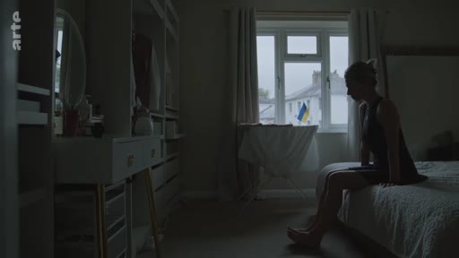 Une femme est assise seule sur son lit dans une chambre sombre, avec un drapeau de l'Ukraine visible à travers la fenêtre en arrière-plan. Cette image illustre le quotidien et les réflexions sur l'identité dans le contexte de la guerre en Ukraine.