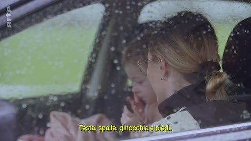 Une femme et un enfant sont assis à l'intérieur d'une voiture alors qu'il pleut à l'extérieur, illustrant le poids psychologique du conflit. Le sous-titre affiche les paroles d'une chanson pour enfants : 'Testa, spalle, ginocchia e piedi'.