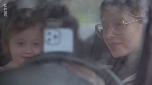 Une femme portant des lunettes et une jeune enfant sont installées à l'intérieur d'une voiture, regardant vers l'avant. Cette scène illustre la vie quotidienne et les moments de complicité familiale dans un contexte potentiellement lié à un déplacement ou à une situation de crise.