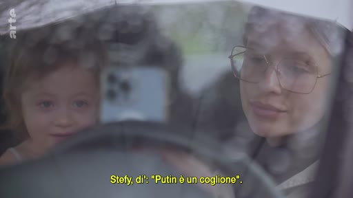 Une femme portant des lunettes filme une jeune enfant dans une voiture, lui demandant de répéter la phrase « Putin è un coglione » (Poutine est un connard), illustrant l'impact psychologique du conflit ukrainien sur les familles.
