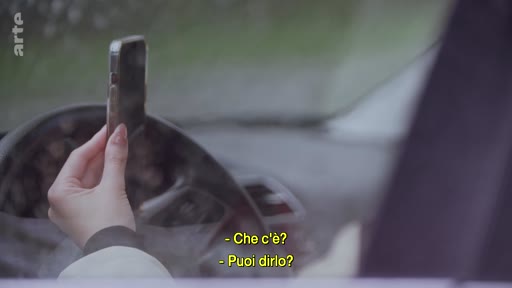 Une personne tient un smartphone à la main tout en étant au volant d'une voiture, illustrant l'utilisation du téléphone au volant. Le sous-titrage en italien affiche « Che c'è? » et « Puoi dirlo? ».