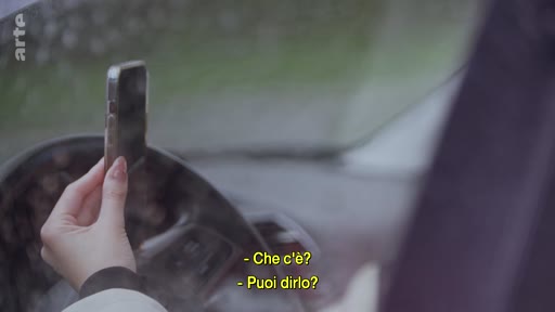 Une personne tient un smartphone à la main tout en étant au volant d'une voiture, illustrant une situation de tension ou de communication lors d'un déplacement.