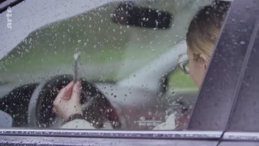 Une conductrice est vue à travers la vitre d'une voiture couverte de gouttes de pluie, illustrant une atmosphère mélancolique ou de tension psychologique.