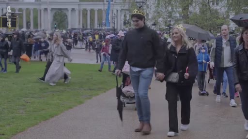 Un couple portant des couronnes en papier marche main dans la main dans le parc près de Buckingham Palace à Londres, au milieu de la foule rassemblée pour le couronnement du roi Charles III. Des passants avec des parapluies circulent dans le parc sous un temps pluvieux.
