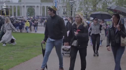 Des membres du public, dont certains portent des couronnes en papier, marchent sous la pluie dans un parc de Londres lors des célébrations du couronnement du roi Charles III. La foule est visible en arrière-plan près d'un monument historique.