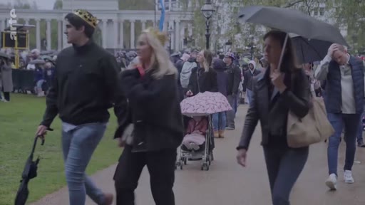 Des membres du public, dont certains portent des couronnes symboliques, marchent dans un parc londonien sous un temps pluvieux lors des célébrations du couronnement du roi Charles III à Londres. La foule est équipée de parapluies et se dirige vers les zones de rassemblement près du palais.