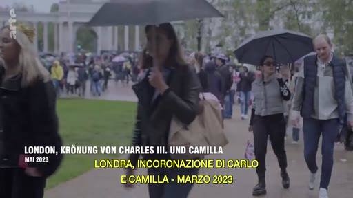 Des passants marchent sous des parapluies à Londres lors des célébrations du couronnement de Charles III et Camilla en mai 2023. La scène montre l'ambiance dans les rues de la capitale britannique.