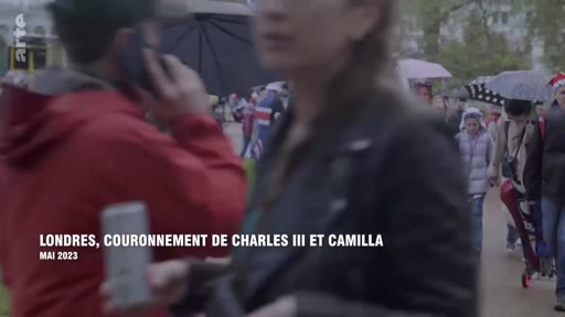 Des spectateurs marchent dans les rues de Londres en mai 2023 lors du couronnement du roi Charles III et de la reine Camilla. La foule est visible avec des parapluies et des drapeaux britanniques.