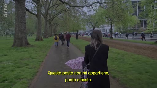 Une femme vue de dos pousse une poussette dans une allée arborée d'un parc public, accompagnée d'un sous-titre en italien sur l'appartenance.