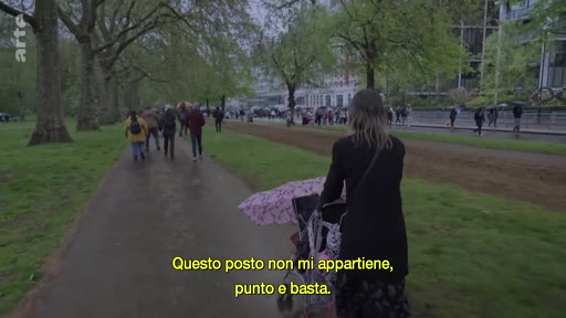 Une femme vue de dos pousse une poussette avec un parapluie ouvert dans une allée de parc, tandis que des passants marchent au loin. Le sous-titre italien exprime un sentiment d'aliénation ou de non-appartenance.