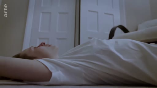 Une personne en t-shirt blanc est allongée sur le sol dans une pièce, les bras étendus au-dessus de la tête, dans une posture évoquant la méditation ou l'introspection.