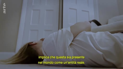 Une personne est allongée sur le sol dans une pièce, accompagnée d'un sous-titre en italien évoquant la présence d'une entité réelle dans le monde.