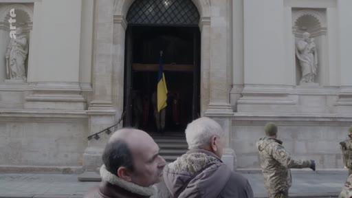 Des civils et des militaires ukrainiens se tiennent devant l'entrée d'une église ornée de statues, où est suspendu un drapeau de l'Ukraine, lors d'une cérémonie funéraire ou commémorative.