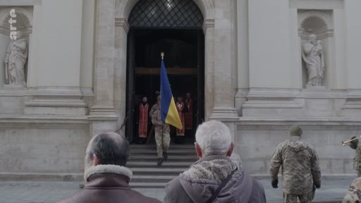 Un soldat en uniforme militaire sort d'une église en portant le drapeau de l'Ukraine, lors d'une cérémonie funéraire ou commémorative. Des personnes assistent à la scène devant l'édifice religieux, illustrant le contexte de la guerre en Ukraine.