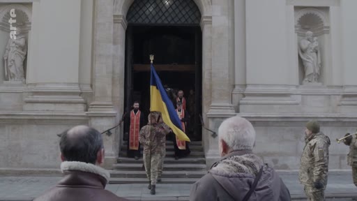 Des soldats en uniforme militaire ukrainien sortent d'une église en portant le drapeau de l'Ukraine lors d'une cérémonie d'hommage funéraire. La scène se déroule devant une façade d'église ornée de statues, illustrant le contexte de la guerre en Ukraine et les funérailles militaires.