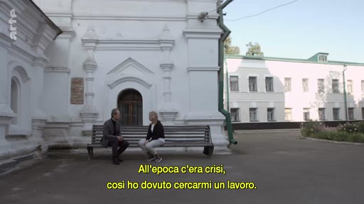 Un homme et une femme sont assis sur un banc en extérieur, devant un bâtiment blanc à l'architecture classique, lors d'une interview. Le sous-titre en italien évoque une période de crise économique et la recherche d'un emploi.