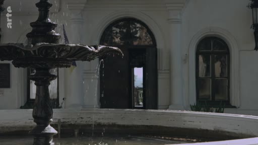 Une fontaine en pierre au premier plan devant l'entrée d'un bâtiment à l'architecture classique, évoquant un cadre propice aux souvenirs d'enfance.
