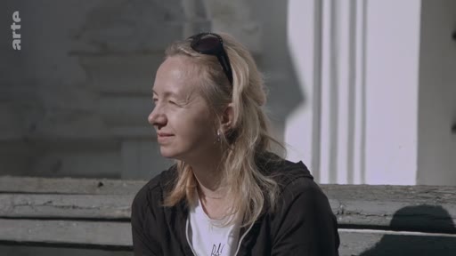 Une femme blonde, portant des lunettes de soleil sur la tête, est filmée en plan moyen lors d'une interview en extérieur, évoquant probablement des souvenirs d'enfance.