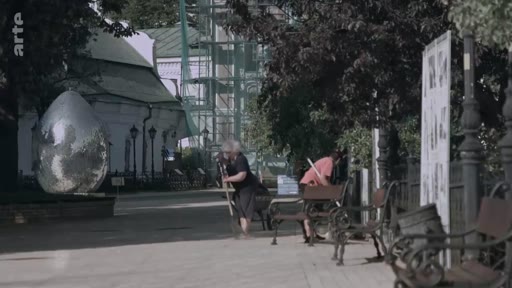 Une employée municipale balaie une allée dans un parc public, à proximité d'une grande sculpture métallique en forme d'œuf et de bancs en bois. La scène se déroule en extérieur dans un environnement urbain ou paysager, avec des travaux de rénovation visibles en arrière-plan.